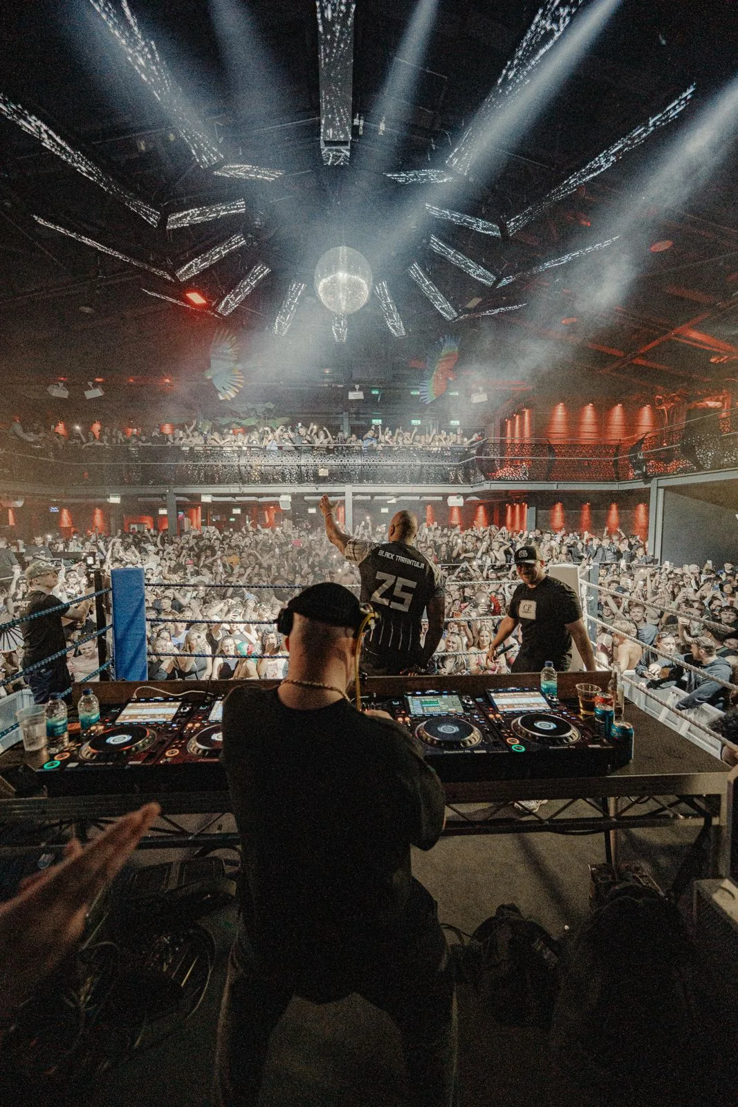 A DJ at the decks seen from behind, facing a packed crowd in a boxing ring stage under a mirror ball and white light beams.